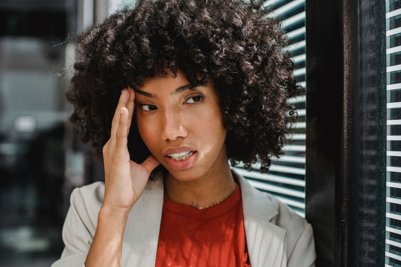 A businesswoman with curly hair expresses stress by her office window, touching her forehead.