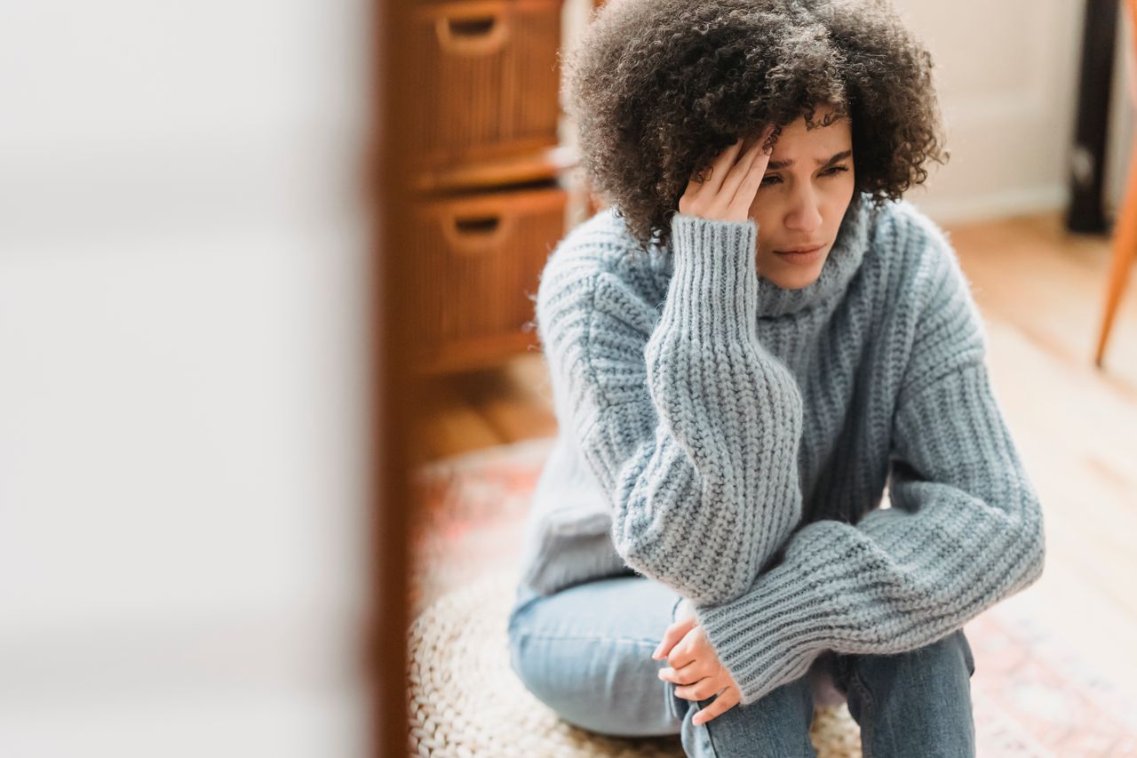 Young ethnic female in casual clothes sitting on pouf while crying with hand on head in light flat