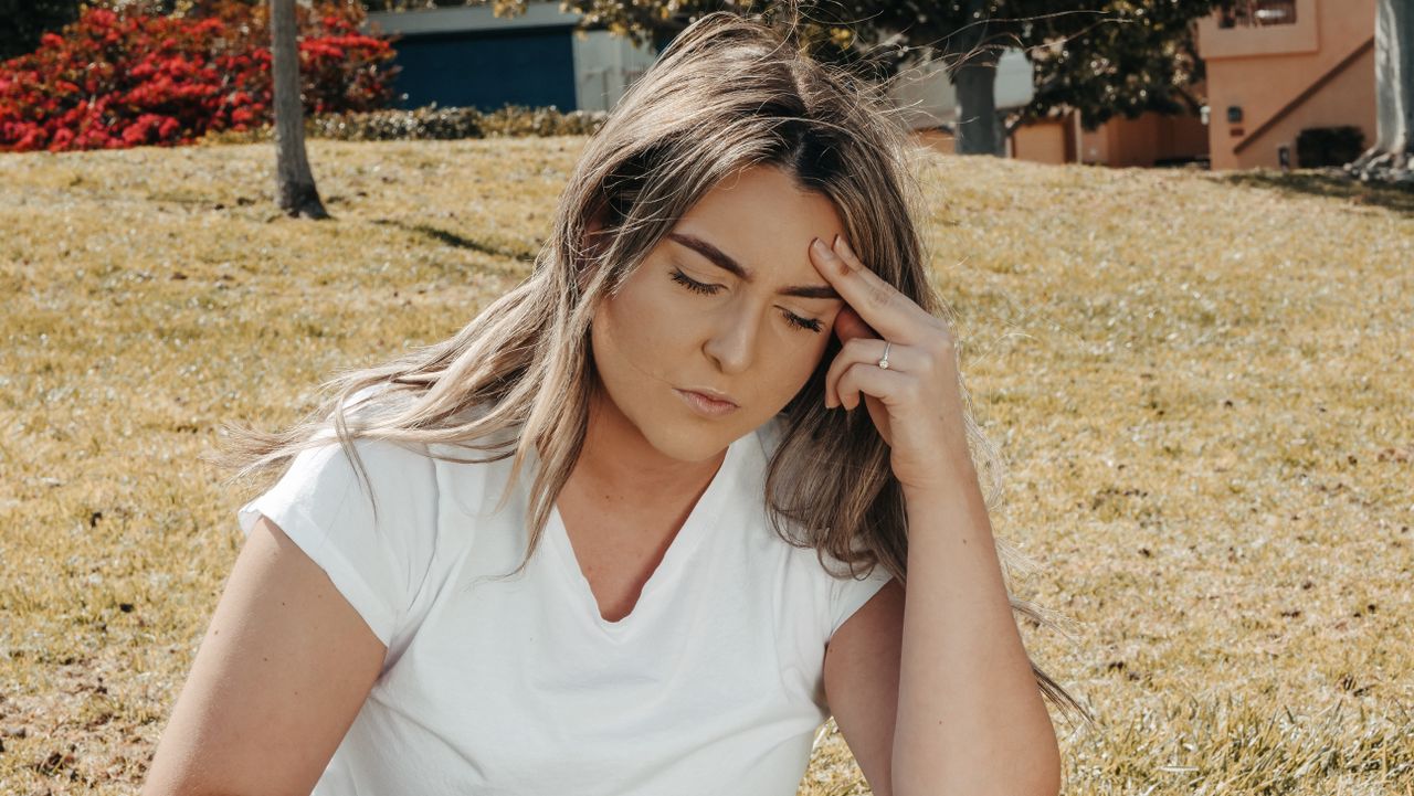 Caucasian woman sitting on grass outdoors with a headache, eyes closed, hand on head.
